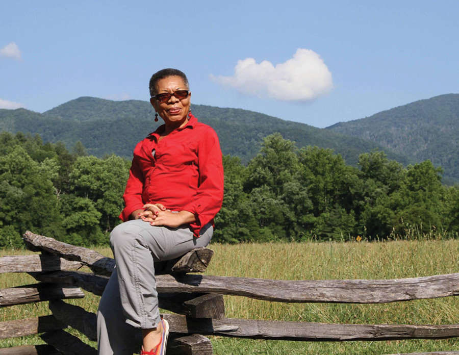 Audrey in a bright red shirt and grey pants sitting on a wooden fence in front of large grasses and hills in the distance