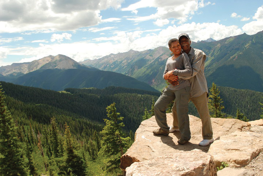 Audrey held by her husband on a large rock at the edge of a deep drop-off that reveals a forest of pine trees at the base and rocky mountains further in the distance