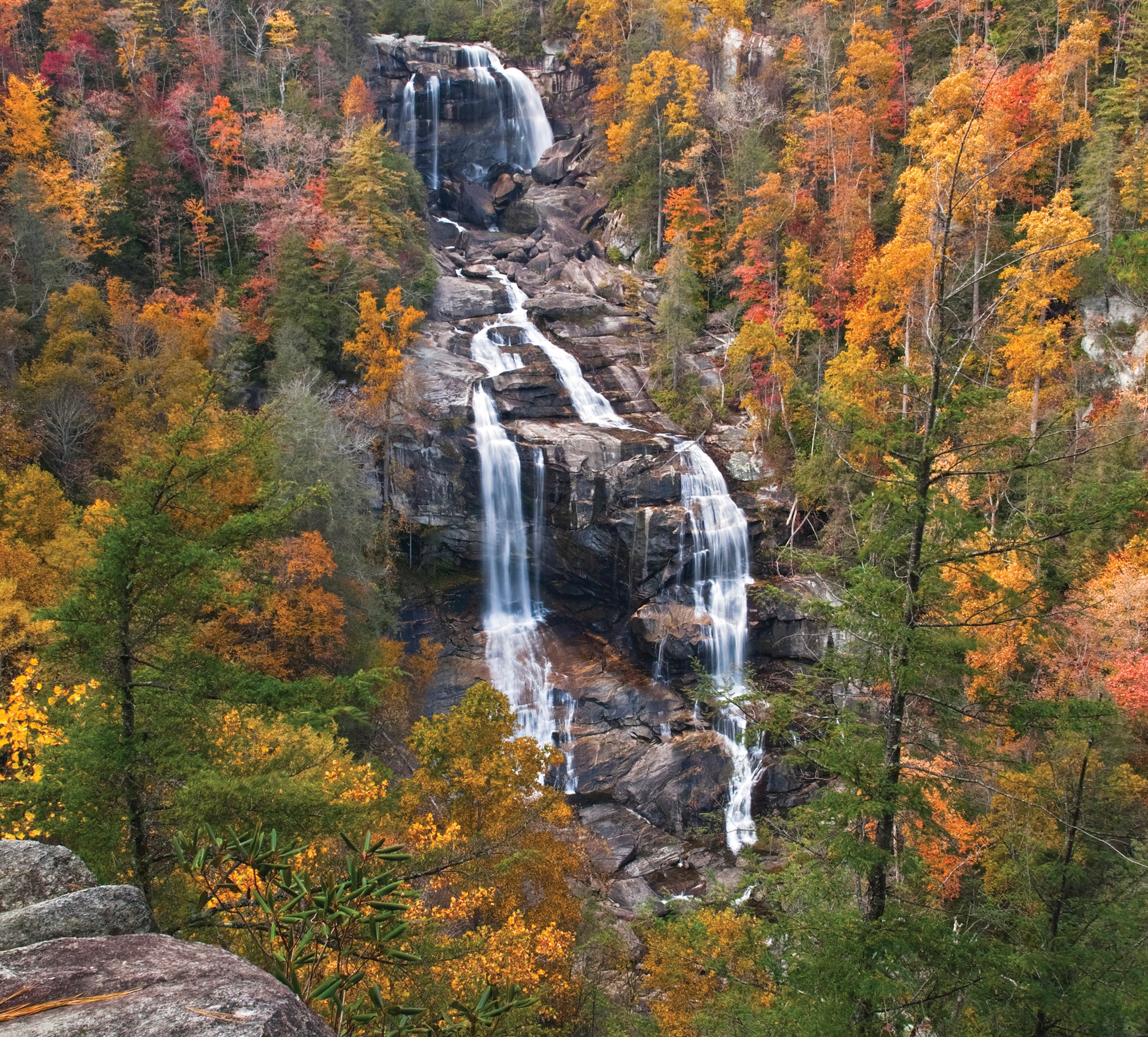 A whitewater waterfall that breaks off into two streams, falling on black rocks in between red,orange, yellow, and green autumn forests