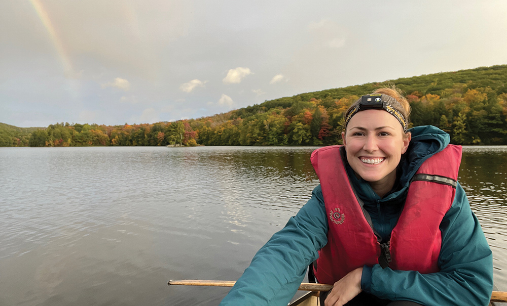 A woman in a aqua blue jacket and red safety vest around her shoulders smiles and poses for a picture in a canoe that floats on top of a lake/river outdoors in the late evening as a rainbow is seen in the far distance