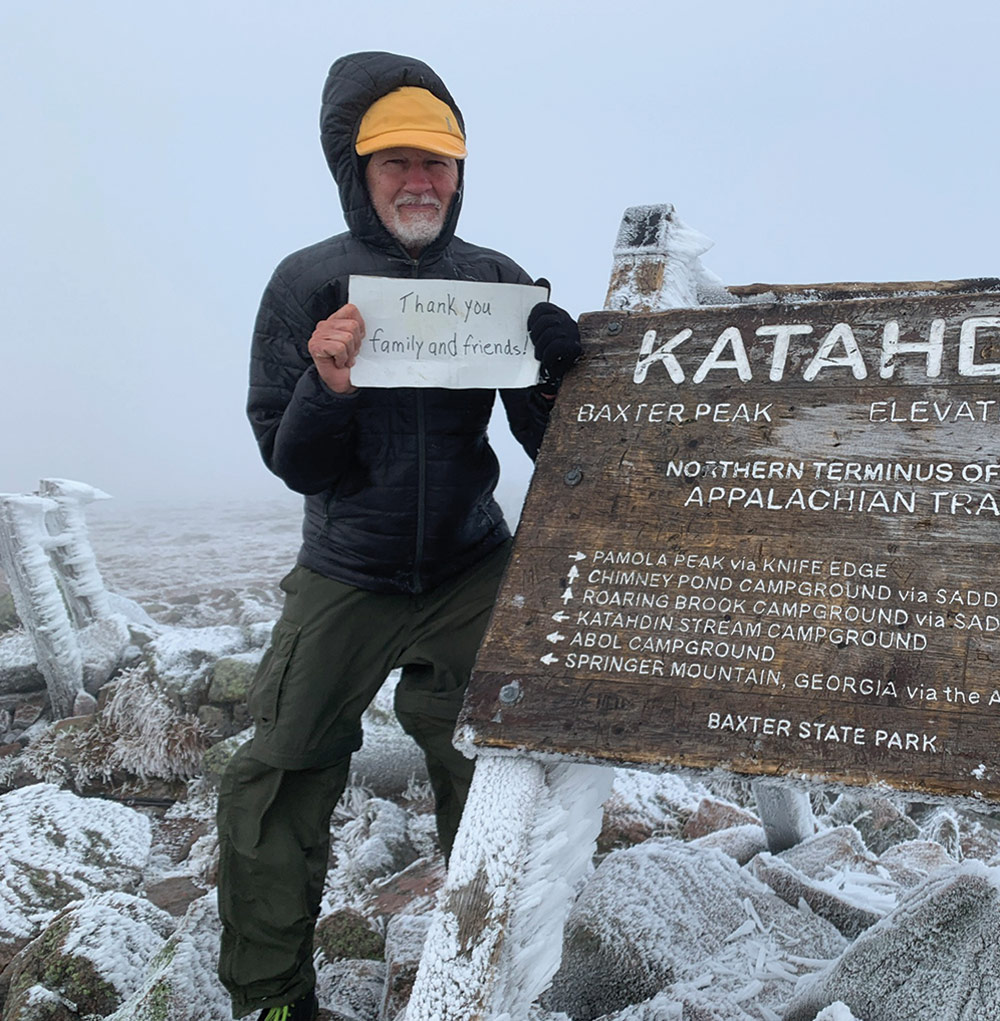 A man in a black puffy jacket and a yellow hat holds a sign that reads "Thank you family and friends" as he stands next to the Baxter Peak wooden post sign outdoors on a snowy overcast day