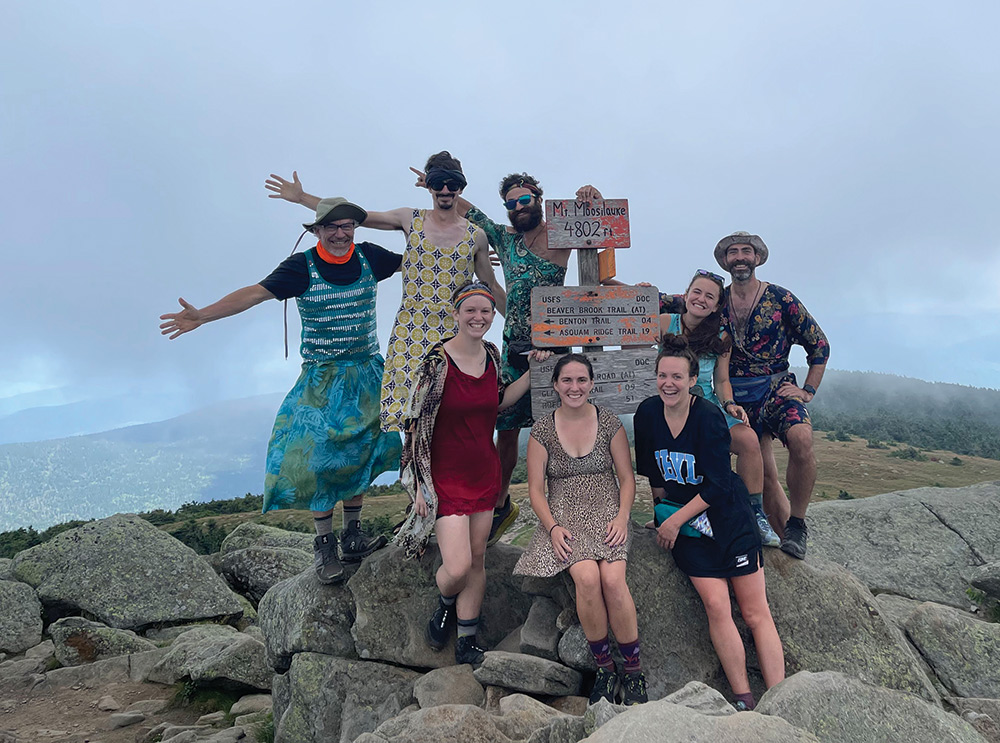 Eight individuals smile and pose for a picture together next to a wooden post sign that reads "Mt. Moosilauke 4802 ft." outdoors on an overcast day