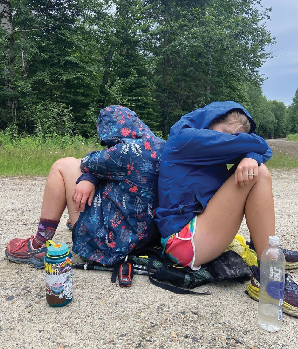 Two individuals are crouched down outdoors on an overcast day in a sitting position with their arms over their knees and hoodies over their heads next to their water bottles nearby a forest area terrain