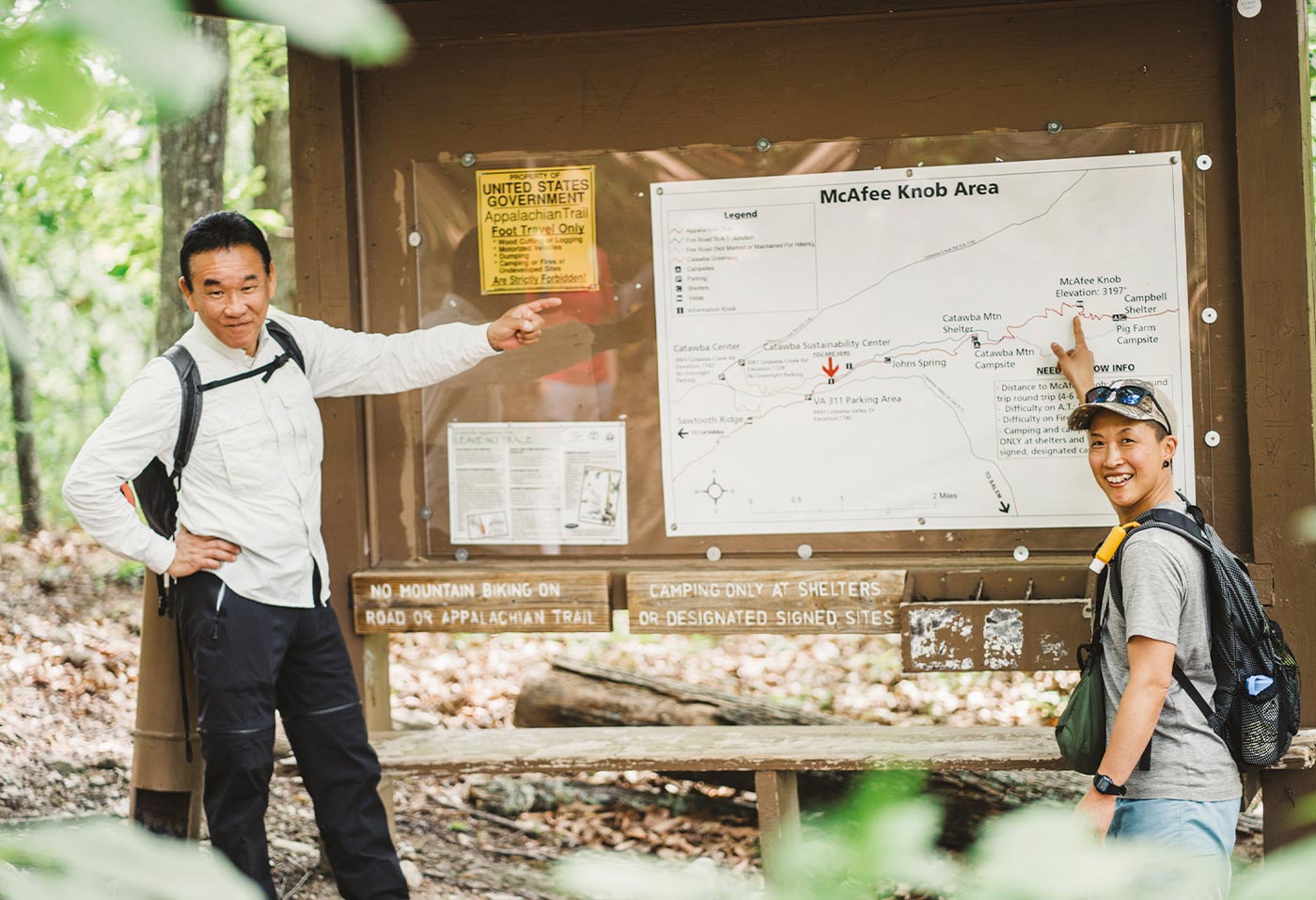 hikers Hiroshi Kimura and Yukiko Satoh stand smiling and wearing backpacks while pointing to the McAfee Knob A.T. trail sign and map