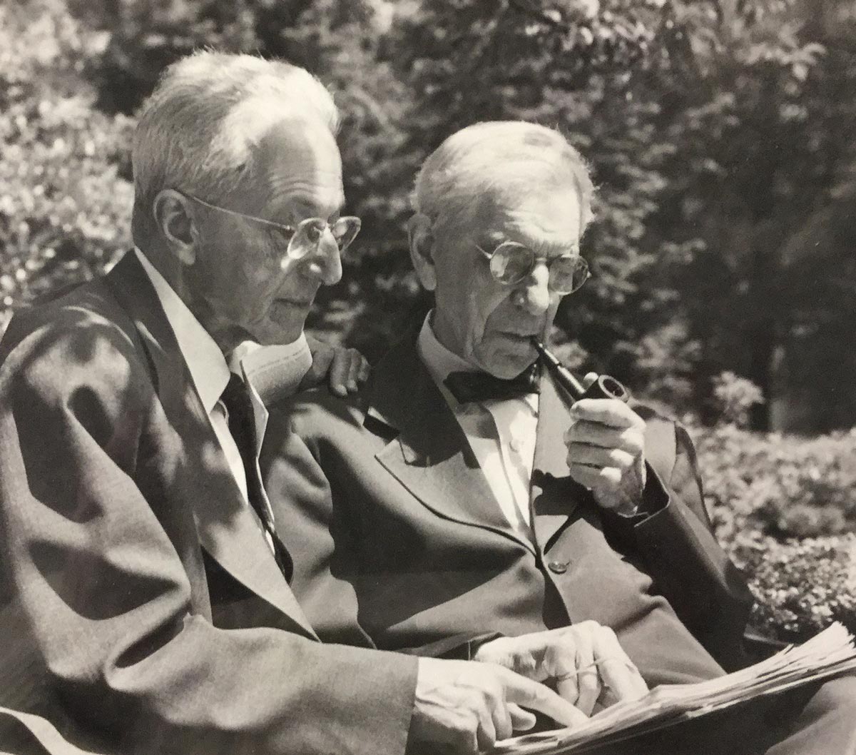 black and white image of two bespectacled men sit outdoors surveying a book together, one of the men smokes a pipe