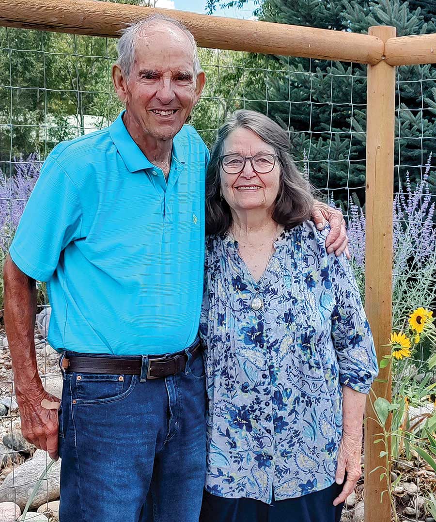 Portrait photograph of Ken and Pat LeRoy smiling together as they pose standing next to each other outside near a wooden fence plus a garden area around them; They live in what they consider “a little slice of heaven,” in Bayfield, Colorado.