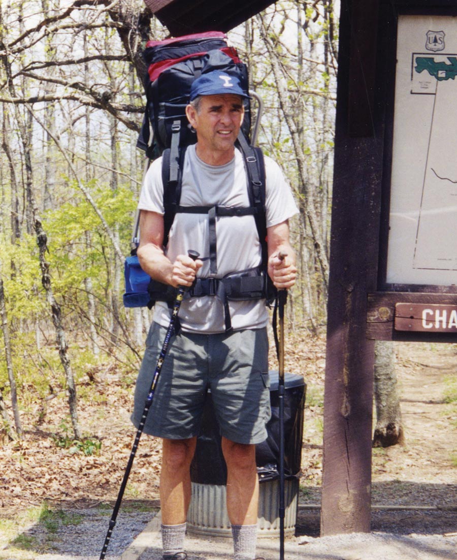 Vintage portrait photograph of Ken LeRoy in hiking gear attire as he has his hiking backpack on plus holding his trekking poles while standing next to a wooden U.S. National Forest Service map sign