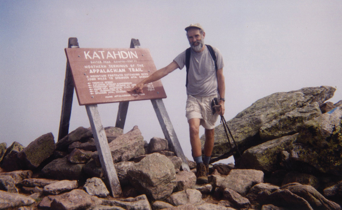 Landscape vintage photograph perspective of Ken LeRoy in hiking gear attire as he is holding his trekking poles and points at the Katahdin Appalachian Trail sign displayed on top of some small and bigger boulders