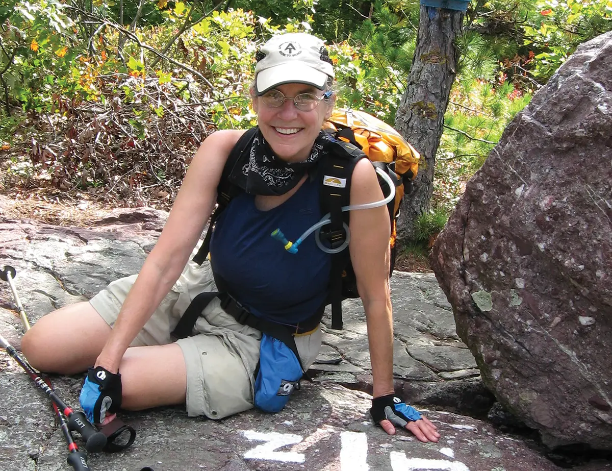Portrait photograph close-up view of Diana Christopulos smiling to take a short pause break at the New York-New Jersey border during one of her northern section hikes as she is seen seated down in casual hiking attire on a rocky ground nearby some grass, tree, and boulder; While dedicated to the protection of the entire A.T., she is especially connected to the part of the Trail near Roanoke, since the McAfee Knob trailhead is about 20 minutes from her home. Christopulos has been hiking that area since 2004 and has patrolled it as a volunteer ridgerunner since 2014, performing light trail maintenance and talking with hikers about the area and how to enjoy the Trail safely and responsibly.