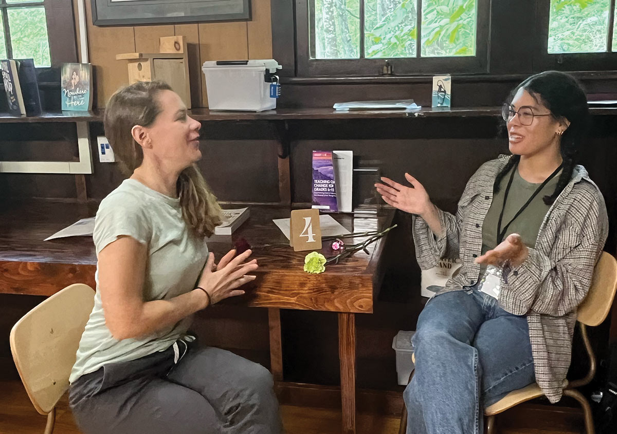 two excited looking young woman engage in a conversation while sitting at a table in a lodge