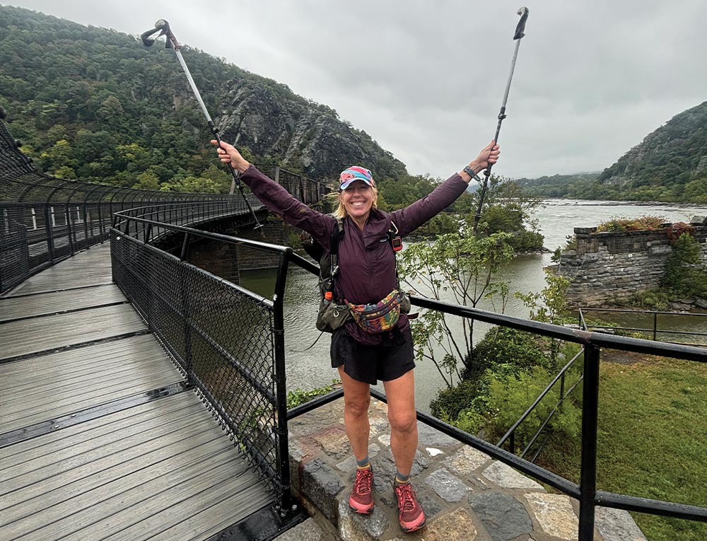 a woman holds her trekking poles aloft and smiles for a picture while standing at the entrance of a partially enclosed bridge overlooking a large creek