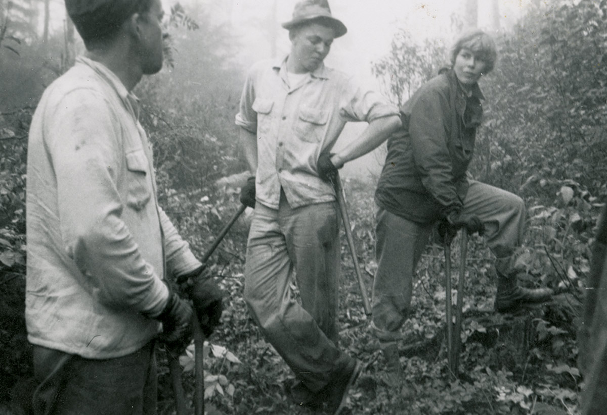 black and white image of three members of a previous outdoor recreation club using work tools in a leafy area