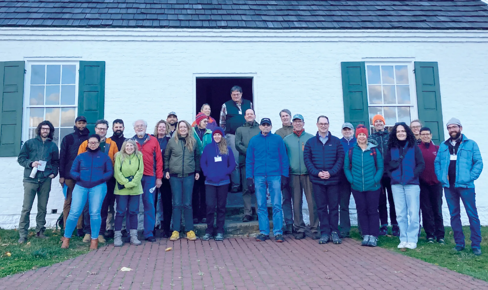 A group of people in outdoor clothing stands in front of a white brick building with green shutters, posing for a group photo on a brick walkway.