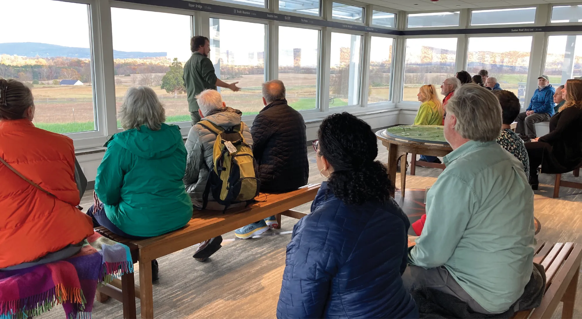 A park ranger stands in front of a large panoramic window, gesturing as he gives a presentation to a group of seated visitors who are looking out at a scenic landscape with farmland and distant hills.
