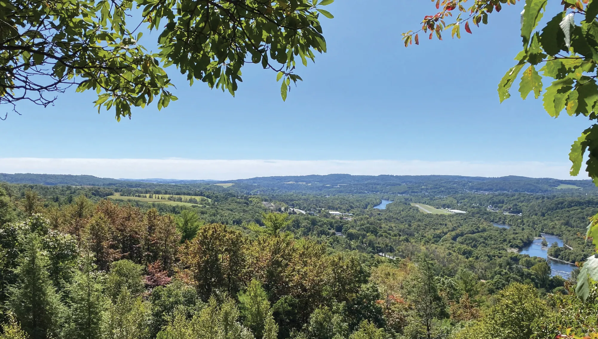 A scenic overlook framed by tree branches shows a winding river cutting through a lush green valley with forests, fields, and low hills under a clear blue sky.