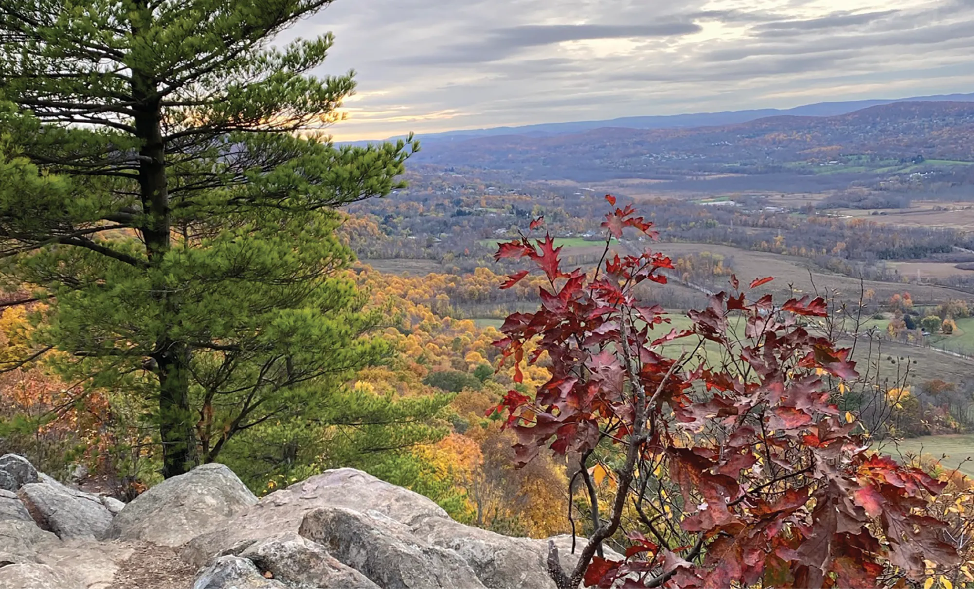 A view from a rocky hilltop with red and green autumn foliage in the foreground, looking out over a patchwork of fall-colored forest and farmland stretching toward distant hills under a cloudy sky.