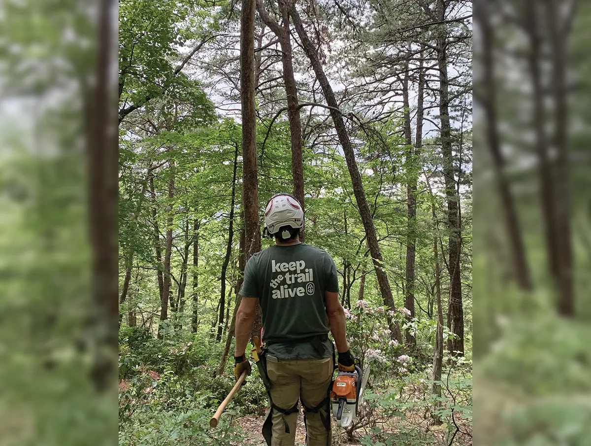 Back view of a trail worker standing in a forest with an axe in one hand and a chainsaw in the other. He wears a white safety helmet and a green T-shirt that reads “Keep the trail alive.” Tall trees and green foliage fill the background.