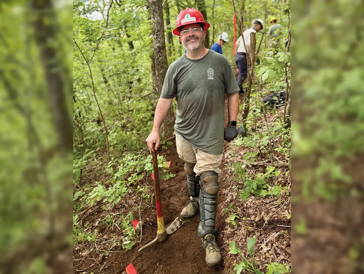 Trail worker in a red hard hat and safety glasses stands on a forest trail holding a tool. He wears a green "STAFF" T-shirt and heavy-duty knee and shin protection. Other workers are seen further back on the trail in the wooded background.