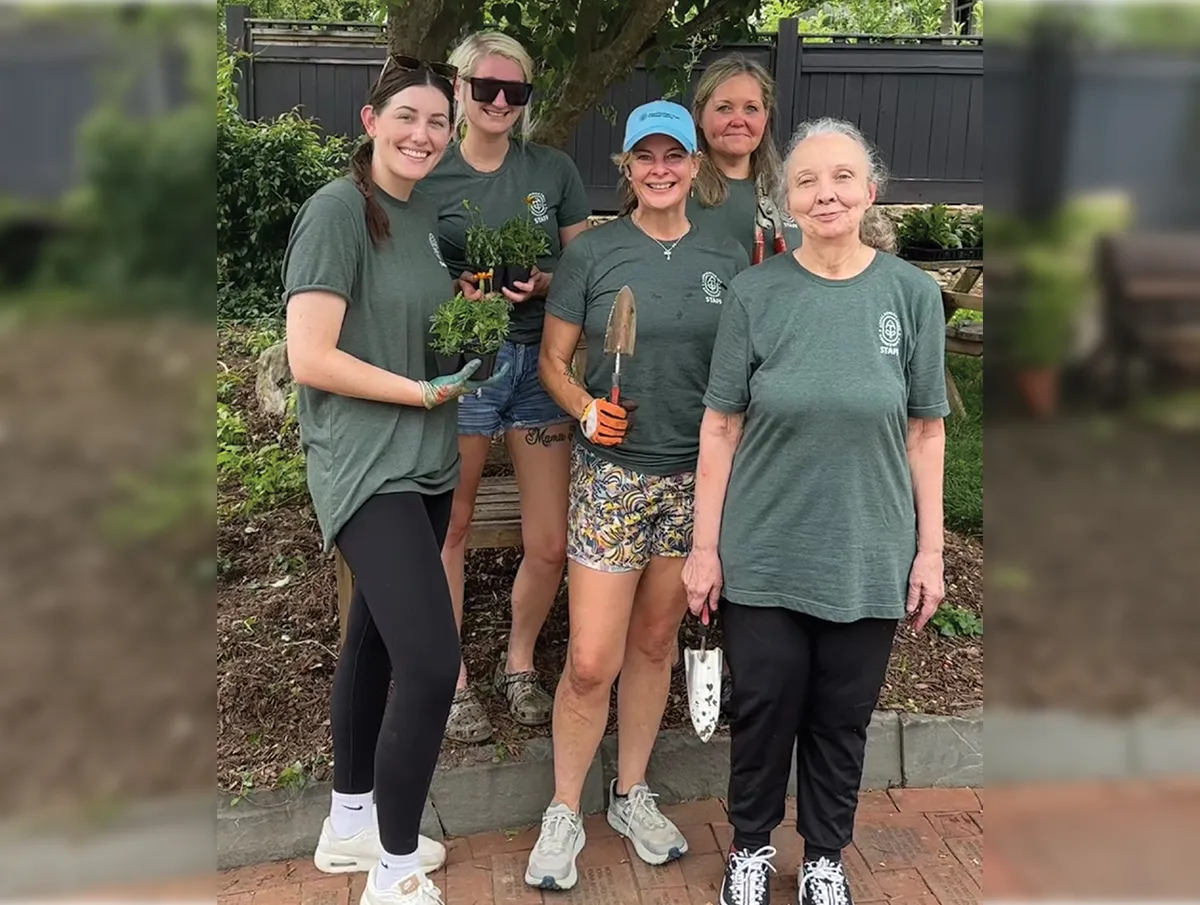 Group of five women standing in a garden area, holding gardening tools and small potted plants. They are wearing matching green staff shirts and smiling at the camera. A dark wooden fence and lush greenery are in the background.