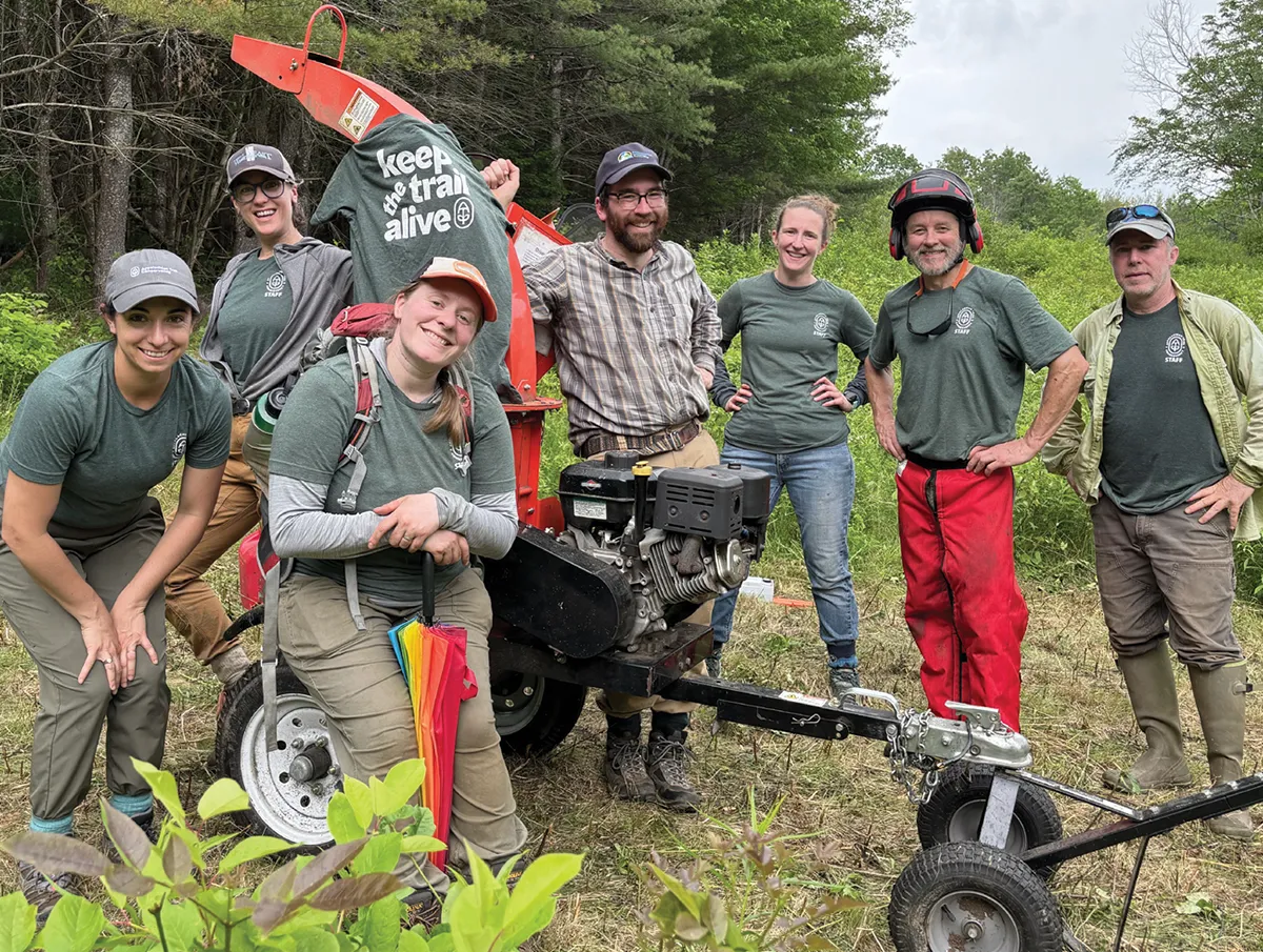 Eight trail staff members gathered around a piece of trail equipment in a clearing surrounded by trees. One shirt draped on the equipment reads “Keep the trail alive.” Everyone is smiling, dressed in work gear, with some holding tools or wearing safety gear.