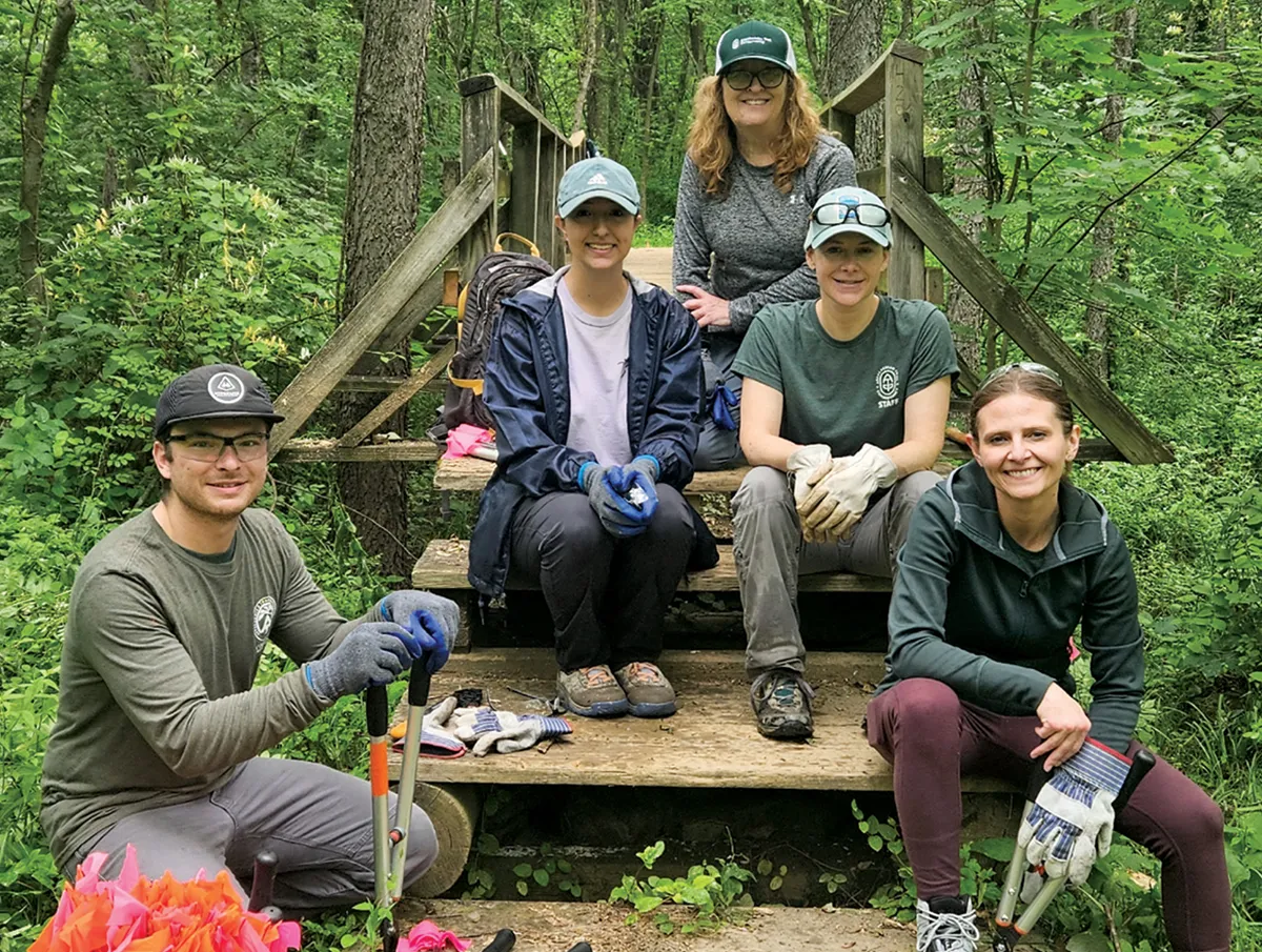 Five people posing on a small wooden footbridge in a forested area. They are wearing trail work clothing including gloves and hats, with tools and equipment placed on the ground nearby. Dense greenery surrounds the scene.