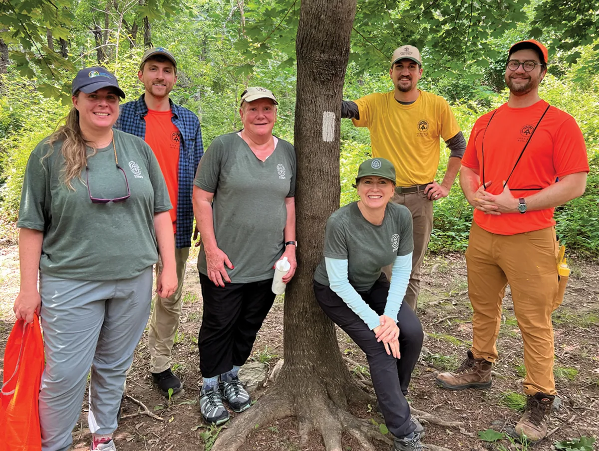 Group of six trail staff members standing in the forest beside a tree marked with a white rectangular trail blaze. They are smiling, wearing outdoor gear including T-shirts and hats, and appear to be in a wooded area with bright green foliage.