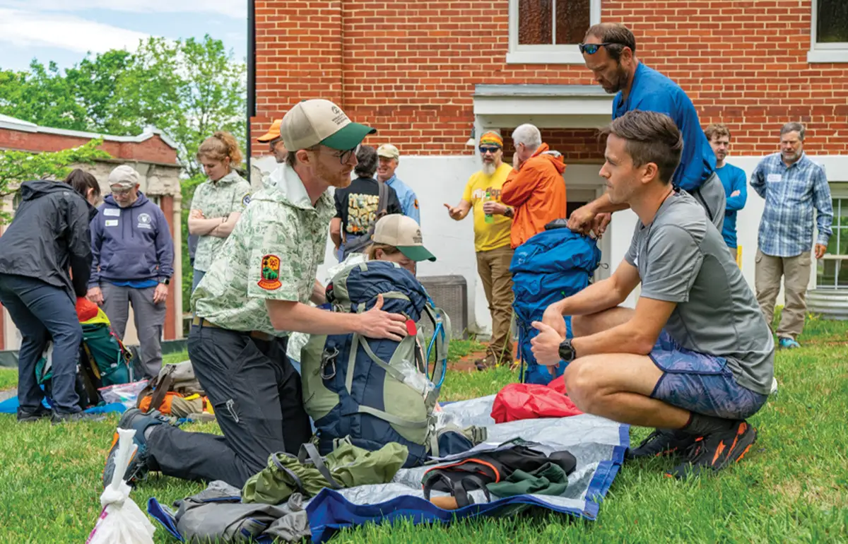 Group of hikers at a trailhead, with two men kneeling to organize or pack their large backpacks and gear on a tarp.
