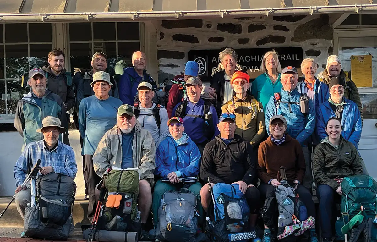 A diverse group of smiling hikers of various ages, many with large backpacks and trekking poles, gathered outdoors in front of a stone building with an 'Appalachian Trail' sign.