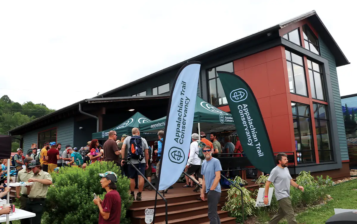 Outdoor event in front of a modern building, with numerous people gathered and prominent "Appalachian Trail Conservancy" banners and tents visible.