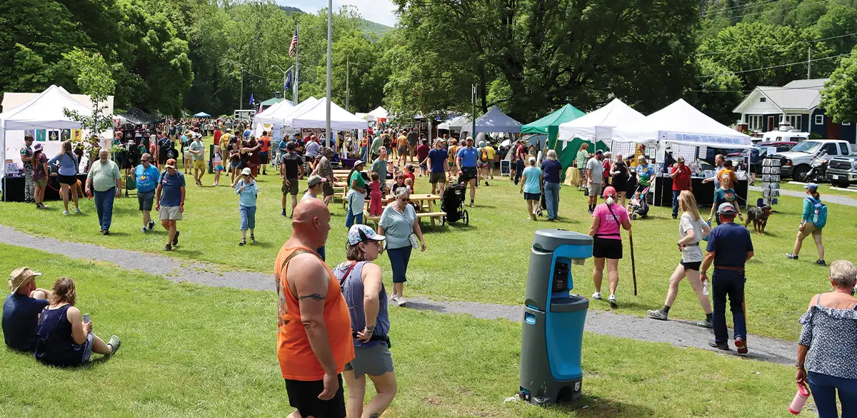 Bustling outdoor festival on a sunny day with numerous people walking among white vendor tents, set in a grassy park area with trees.