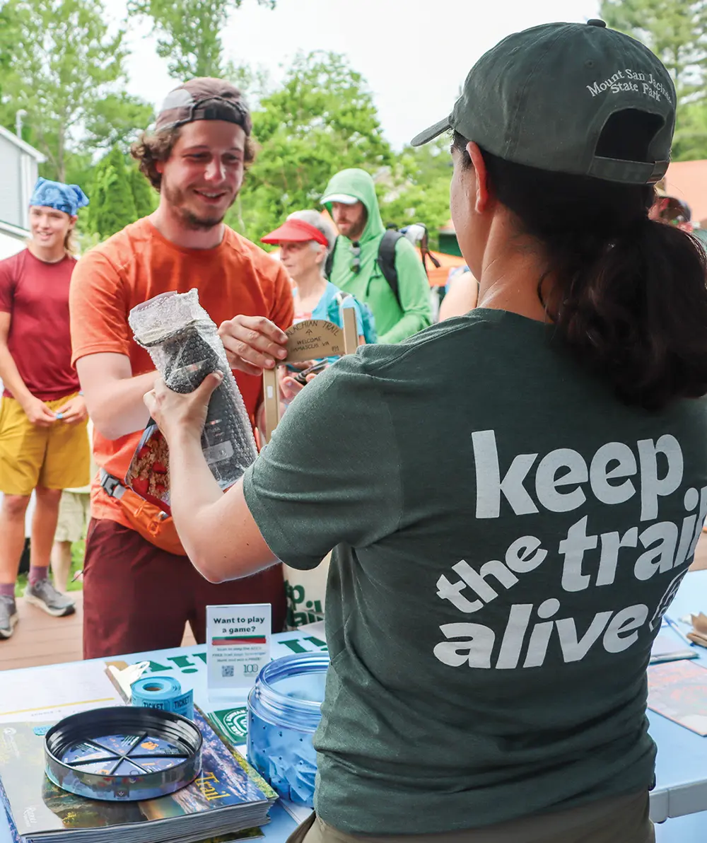 A volunteer wearing a "keep the trail alive" t-shirt handing an item to a smiling male hiker at an outdoor information booth