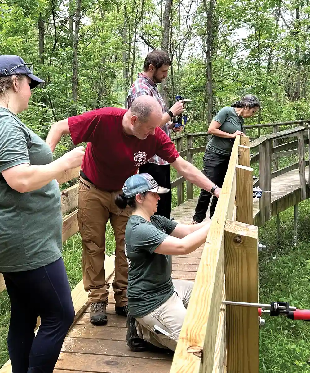 A group of volunteers in a wooded area working together to construct a wooden bridge, using tools to secure railings.