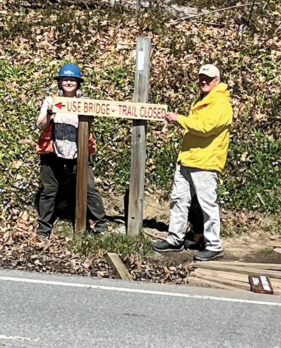 Two people holding a ‘Use Bridge - Trail Closed’ sign.