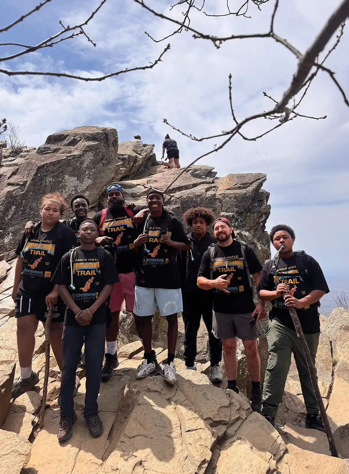 Group of diverse young hikers on a rocky mountain summit wearing matching shirts.