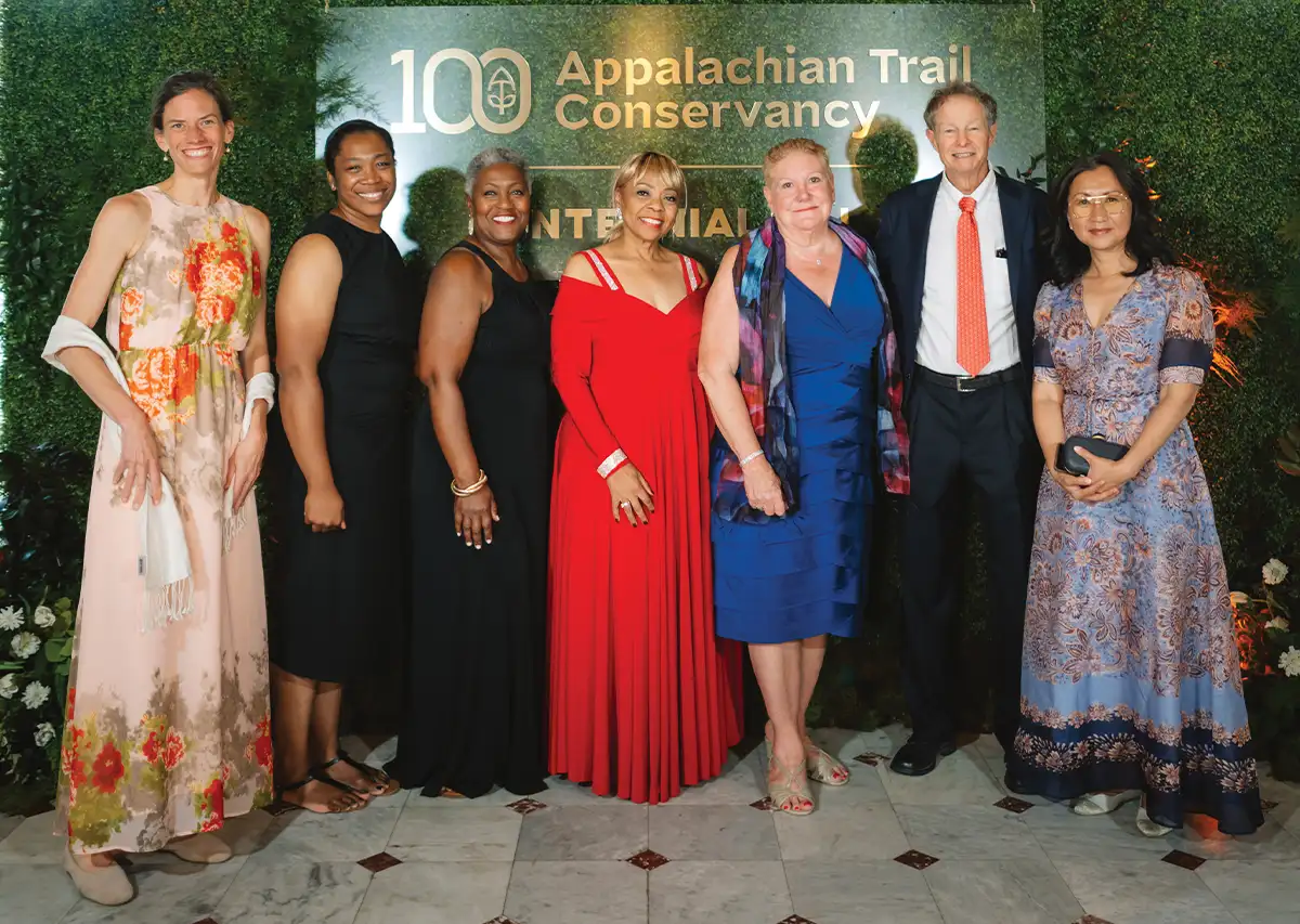 Seven smiling individuals, formally dressed, posing in front of a backdrop with the ‘100 Appalachian Trail Conservancy Centennial’ logo.