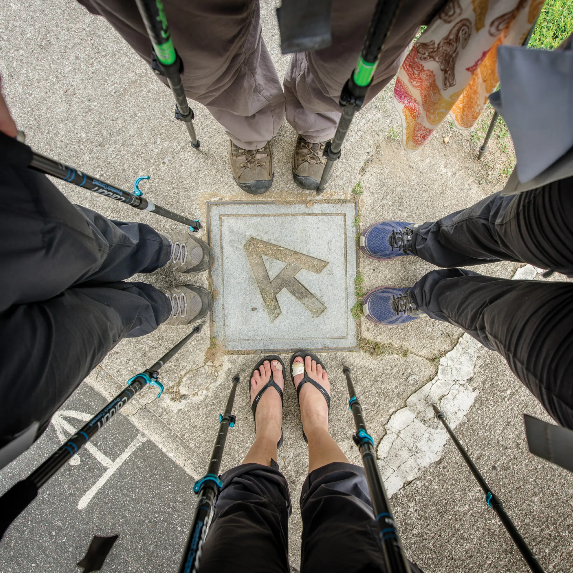 Overhead view of five hikers standing in a circle around a concrete Appalachian Trail marker embedded in the pavement, each holding trekking poles and wearing different types of footwear, including hiking boots, sneakers, and flip-flops.