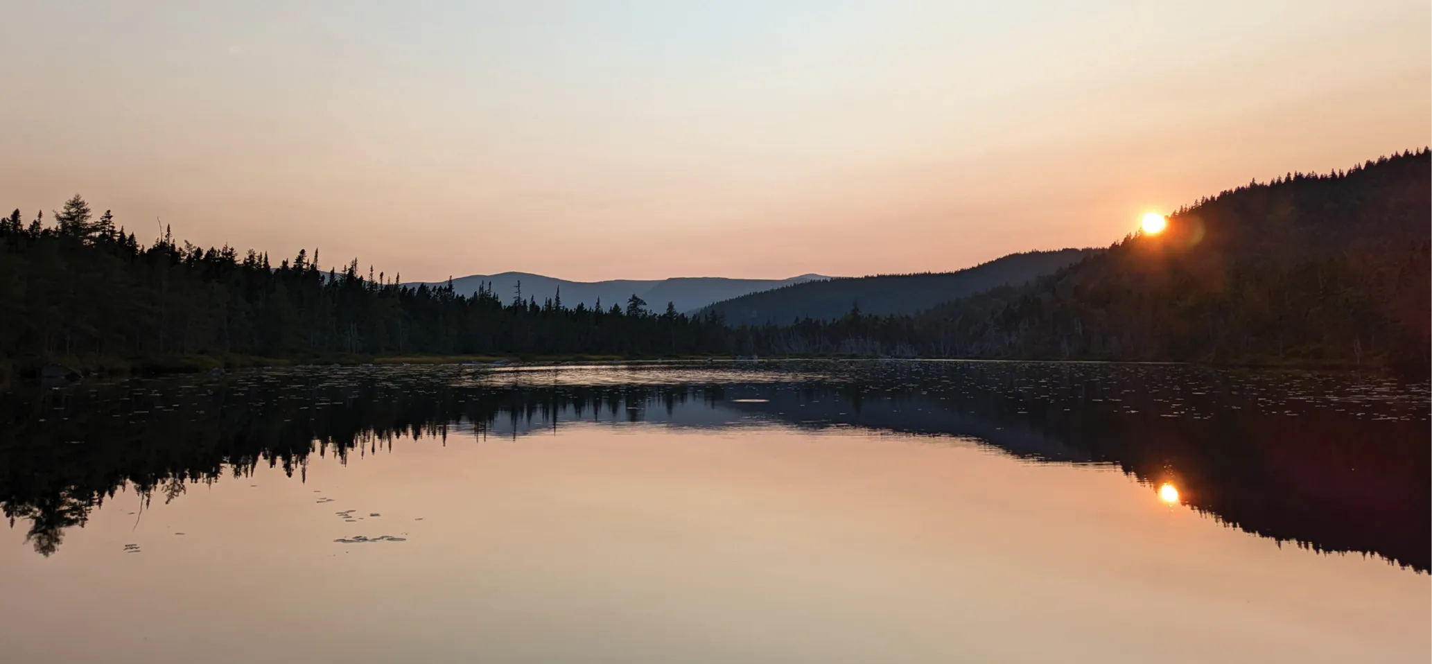Serene lake scene at sunset with the sun dipping behind a forested ridge, casting a golden reflection across the still water under a soft pink and orange sky.