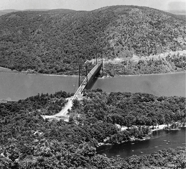 Historic black-and-white aerial photo of a suspension bridge spanning a wide river, with wooded hills rising steeply on both sides.