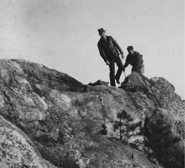 Black-and-white photo of two hikers climbing across rocky terrain on a steep slope.