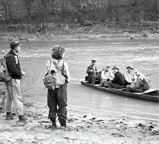 Black-and-white photo of hikers standing on a riverbank while a group of men in a wooden boat prepare to ferry them across.