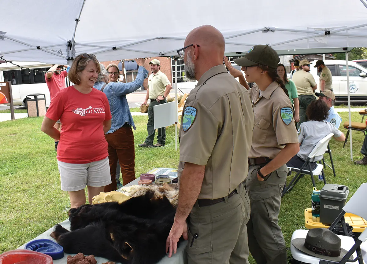 Park rangers in uniform talking to visitors at an outdoor outreach booth displaying black bear pelts and natural history items.