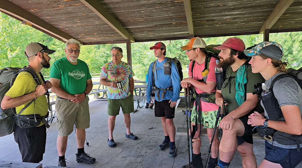A mix of six hikers, some heavily packed, and two casual visitors talk under a wooden pavilion roof on a sunny day.