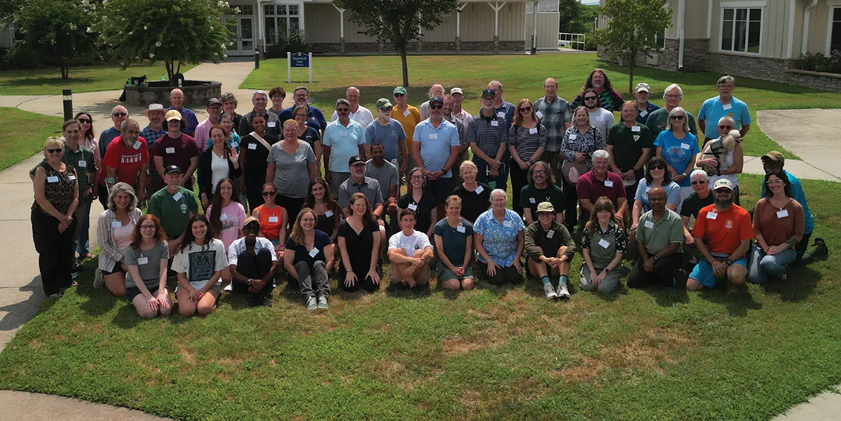 A large group of approximately fifty people, of varying ages and genders, poses outdoors for a group photo on a sunny day on a green lawn.