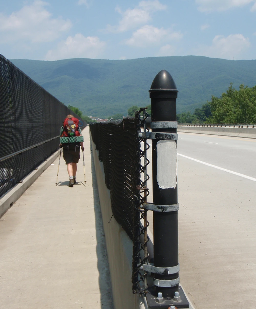 A hiker with a large red backpack and trekking poles walks away from the camera on the concrete shoulder of a highway bridge or overpass. A chain-link fence separates them from the highway, and lush green mountains are visible in the background. A black pole in the foreground has a white blaze painted on it.