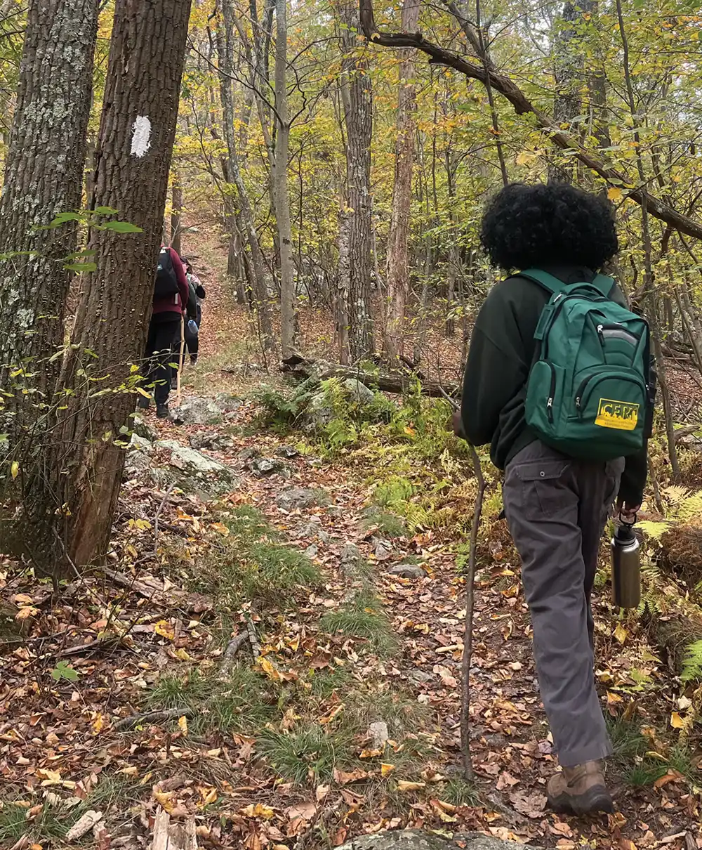 A photo of hikers on a rocky, leaf-covered trail in a lightly forested area during the fall. A prominent white trail blaze is visible on a tree trunk in the foreground on the left, as a hiker with a large, green backpack walks away from the camera.