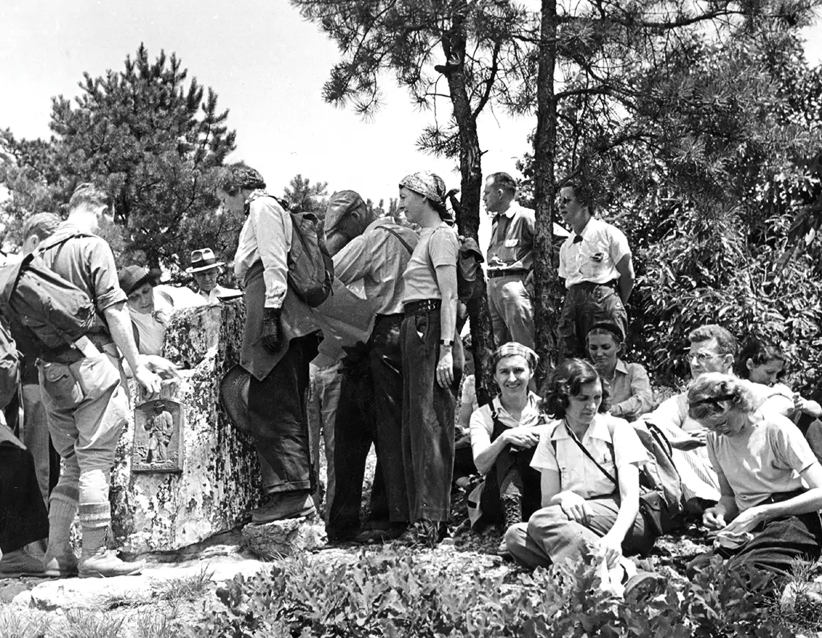 A black and white historical photo of a group of hikers, dressed in 1930s or 1940s clothing, gathered on a rocky outcrop. Several are standing and looking at a marker or plaque, while others are seated in the foreground.