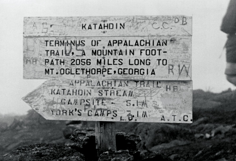 A grainy, black and white historical photo of a wooden trail sign, covered in scratched graffiti. The main text reads, "KATAHDIN TERMINUS OF APPALACHIAN TRAIL, A MOUNTAIN FOOT-PATH 2056 MILES LONG TO MT. OGLETHORPE, GEORGIA."