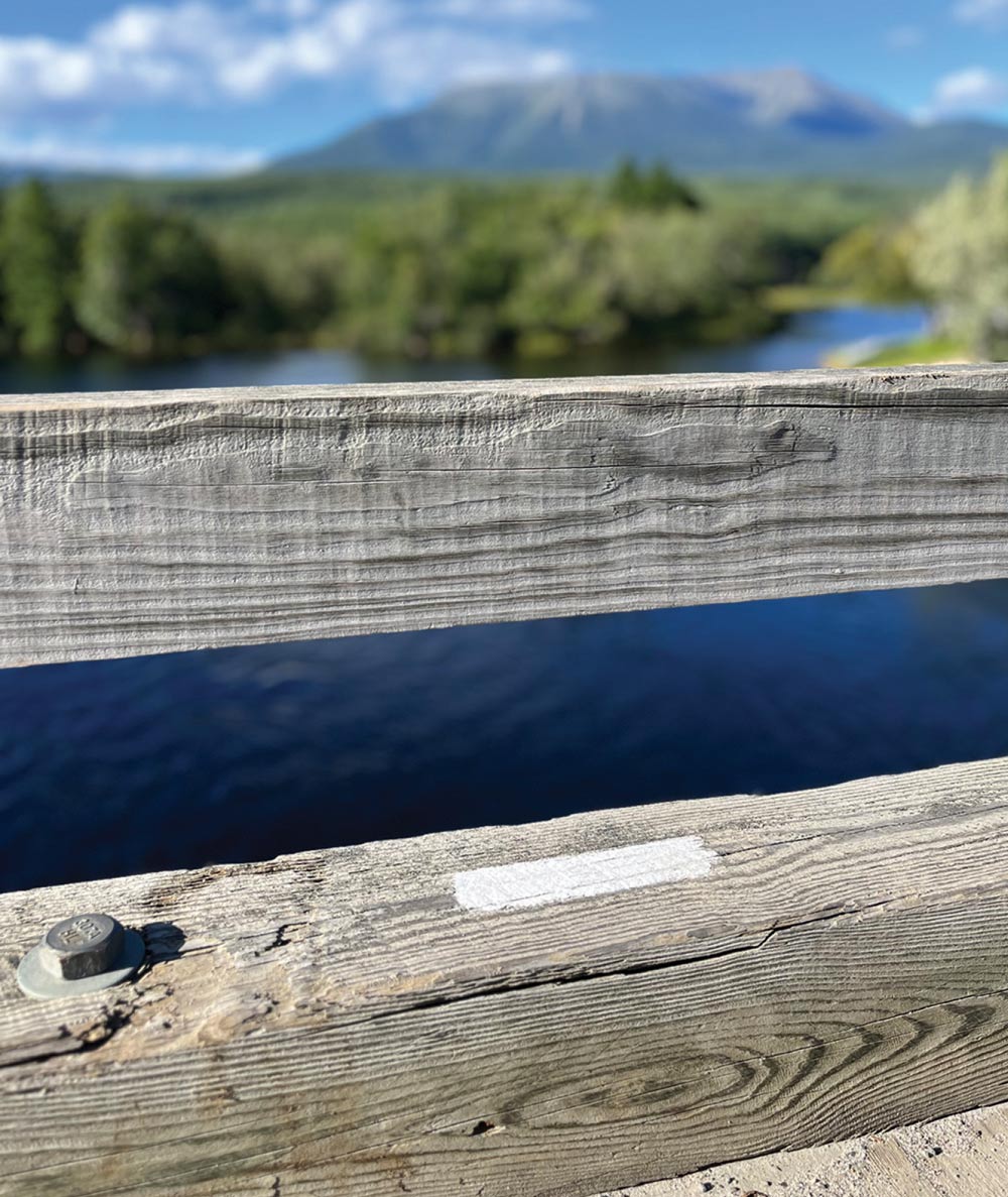 A close-up photo of a wooden bridge railing with a bright white trail blaze painted on the horizontal rail near a bolt. In the blurry background, a dark blue river or lake is visible with a tree line and a large, distant mountain (likely Katahdin).