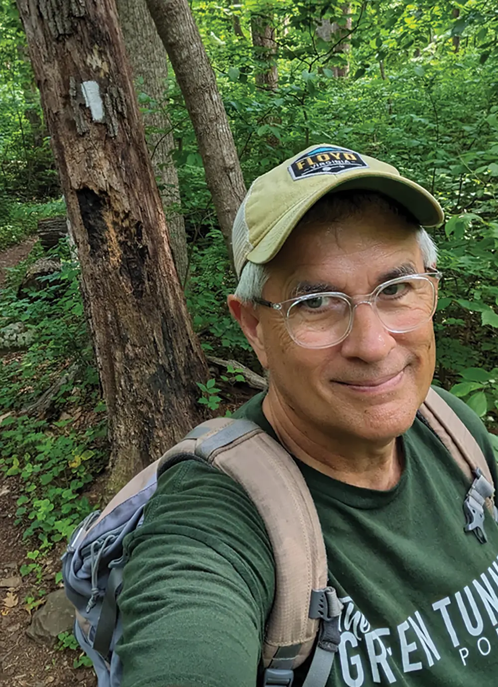 Mills Kelly smiling, wearing a green t-shirt and baseball cap, standing next to a tree with a white trail blaze. He has a backpack on and is surrounded by dense green forest foliage.