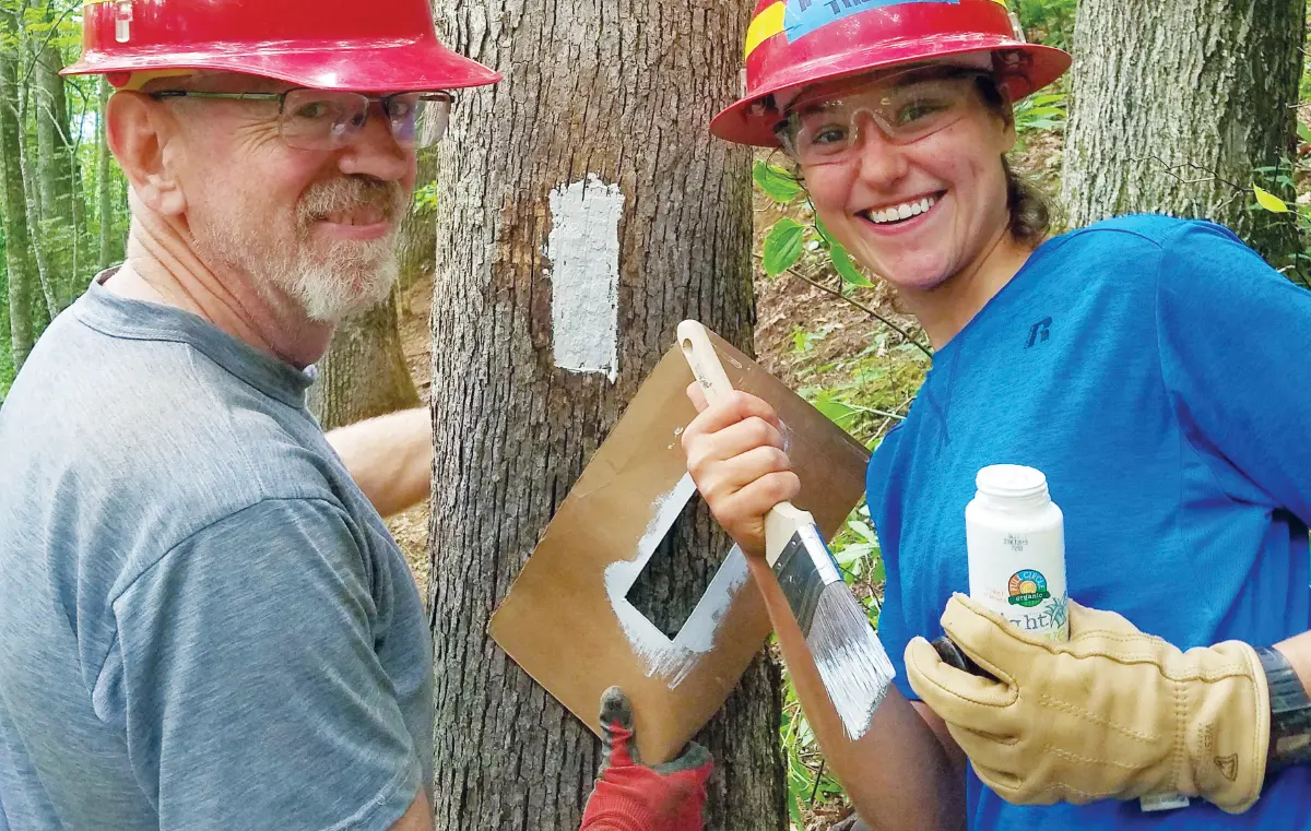 Two trail maintenance volunteers, a man and a woman, are smiling while painting blazes on a tree; Both are wearing red hard hats and the woman is wearing safety glasses; They are using a brown paper diamond-shaped stencil to paint white trail markers (blazes) onto the tree; The woman holds a can of paint and a paintbrush